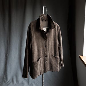 May include: A brown button-down jacket with two front pockets hangs on a metal hanger. The jacket has a wide collar and long sleeves. The fabric appears to be a textured wool or wool blend. The jacket is displayed against a dark gray backdrop.