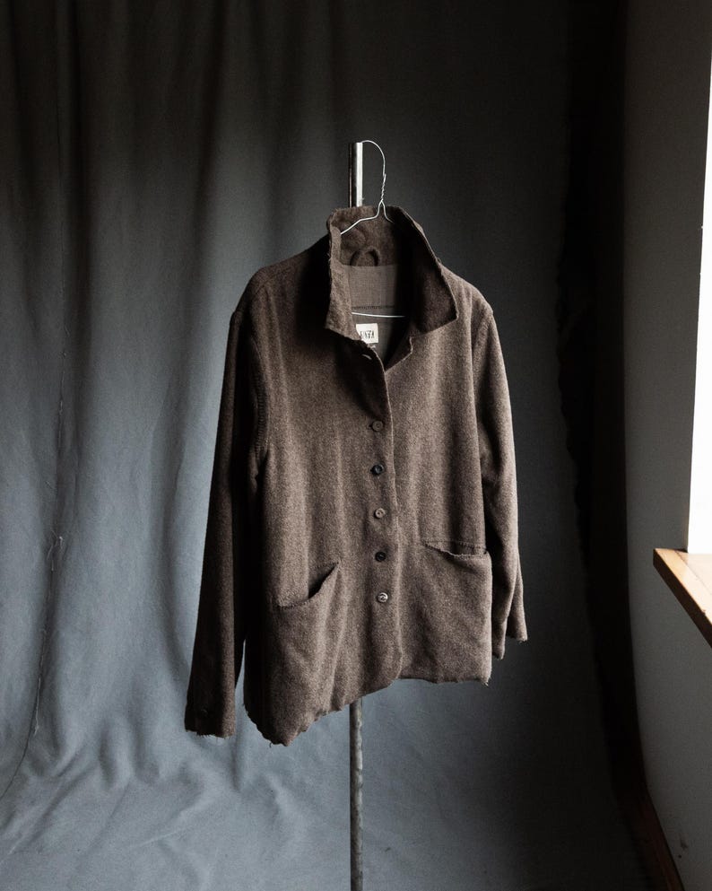 May include: A brown button-down jacket with two front pockets hangs on a metal hanger. The jacket has a wide collar and long sleeves. The fabric appears to be a textured wool or wool blend. The jacket is displayed against a dark gray backdrop.