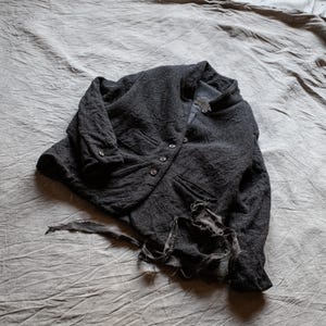 Puede incluir: Una chaqueta gris carbón oscuro con una superficie texturizada y rugosa, que presenta un cierre frontal de botones, cuello alzado, mangas largas con puños abotonados y un dobladillo deshilachado con tiras de tela colgando del borde.