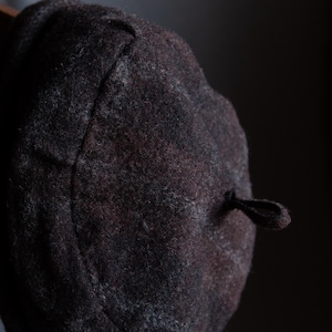 May include: A dark grey wool beret with a black cord and tassel. The beret has a subtle plaid pattern.