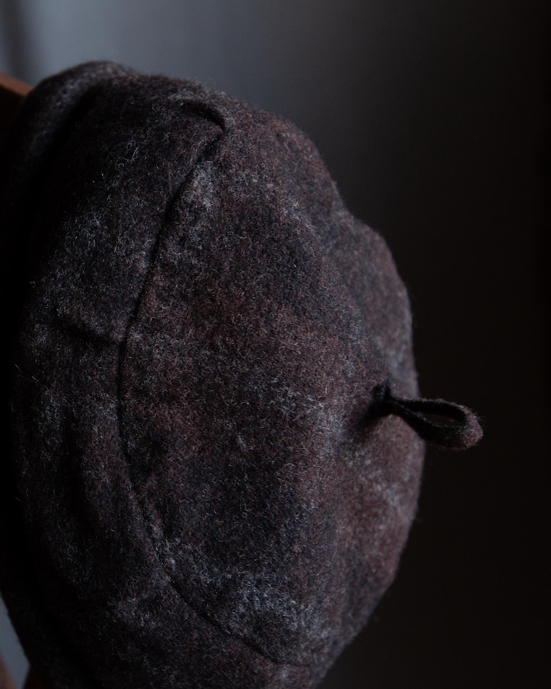 May include: A dark grey wool beret with a black cord and tassel. The beret has a subtle plaid pattern.