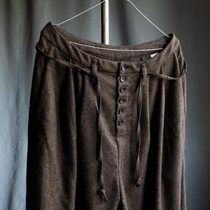 Puede incluir: Pantalones marrones con estampado de espiga colgados de una percha de alambre. Los pantalones tienen cintura alta, bragueta abotonada y detalles de cordón. La tela parece ser de peso medio. Los pantalones son de color neutro.