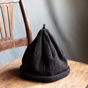 Könnte beinhalten: Eine dunkelbraune Beanie-Mütze mit einer abgerundeten Krone und einem kleinen, geknoteten Detail oben. Die Mütze ist aus einem strukturierten Stoff gefertigt und liegt auf einem Holzstuhl. Die Farbe der Mütze hat rote und graue Flecken.