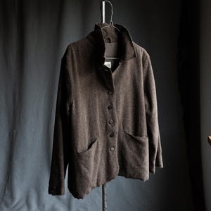 May include: A brown jacket with a high collar and two front pockets hangs on a metal hanger. The jacket has a button-up front and a textured appearance. The background is a dark gray.