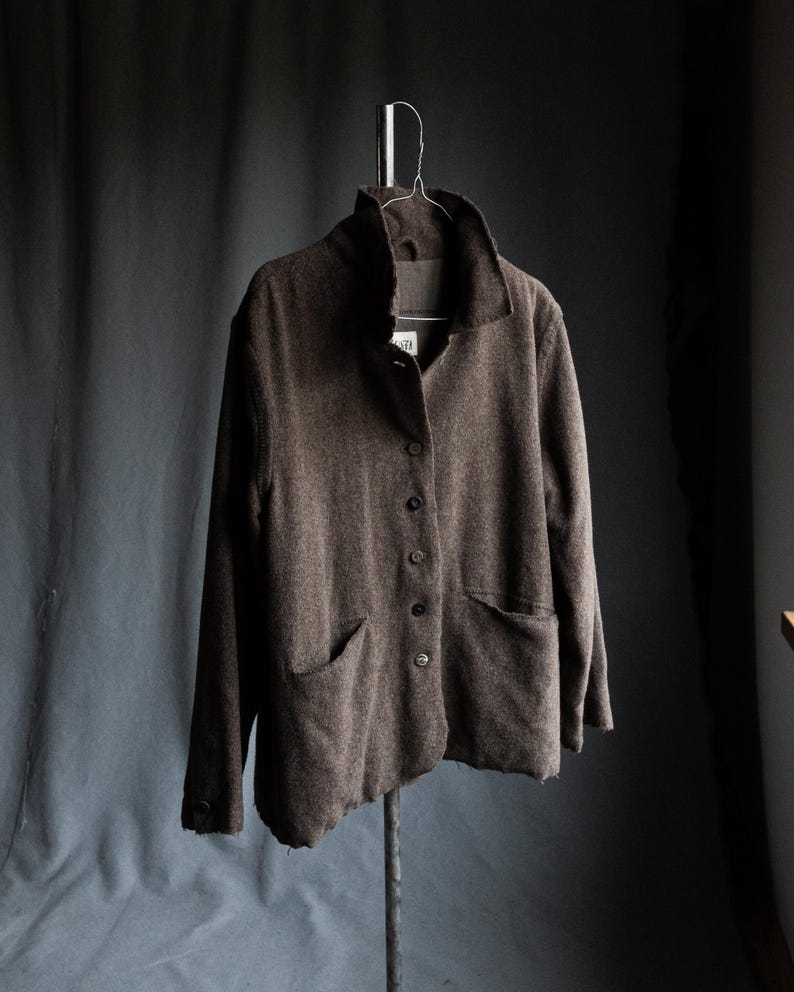 May include: A brown jacket with a high collar and two front pockets hangs on a metal hanger. The jacket has a button-up front and a textured appearance. The background is a dark gray.