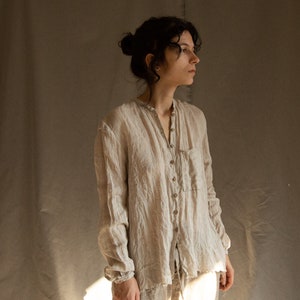 Può includere: Una donna indossa una camicia di lino beige chiaro con bottoni sul davanti e una singola tasca sul lato sinistro. La camicia ha una vestibilità rilassata e un orlo leggermente sfrangiato.