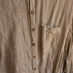 Può includere: Una camicia di lino color marrone chiaro con chiusura a bottoni sul davanti e una sola tasca sul lato sinistro. La camicia ha una vestibilità rilassata e un aspetto leggermente usurato.