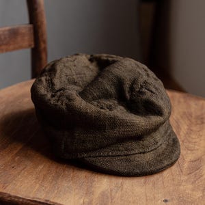 May include: A brown newsboy cap made of textured fabric sits on a wooden chair. The cap has a rounded crown and a short, stiff brim. The color is a deep, earthy brown, and the fabric appears to be woven.