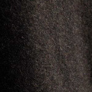 May include: Close-up of a dark brown, textured fabric. The material appears to be a dense wool or similar textile, with a slightly uneven surface and visible fibers. The color is a deep, rich brown.