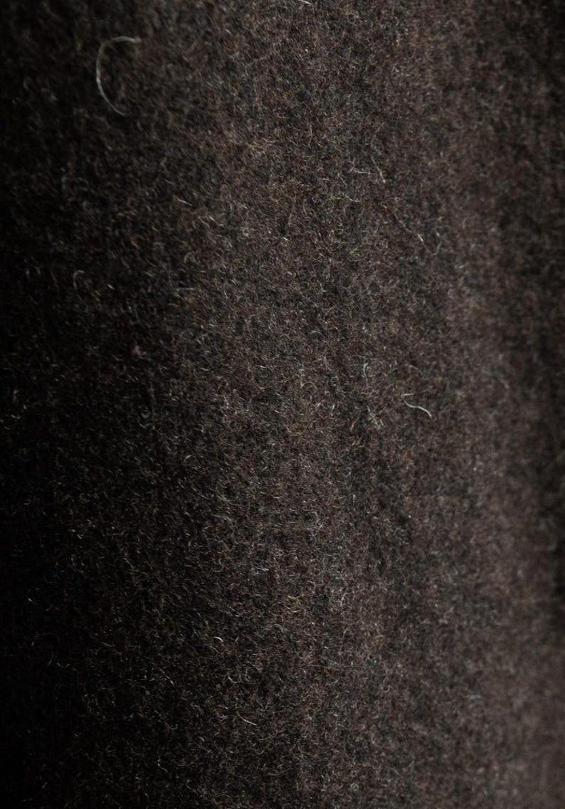 May include: Close-up of a dark brown, textured fabric. The material appears to be a dense wool or similar textile, with a slightly uneven surface and visible fibers. The color is a deep, rich brown.