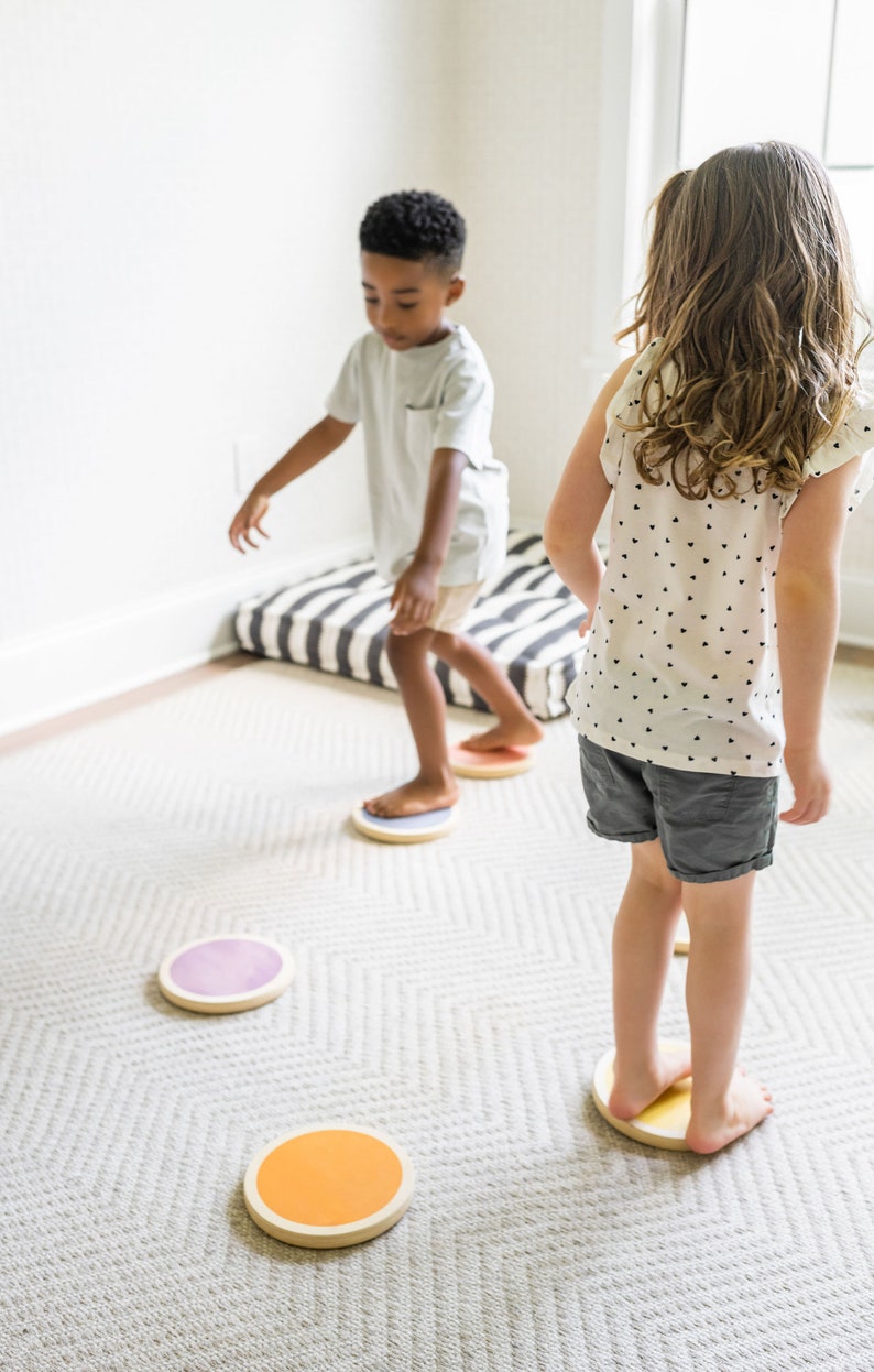 A dark-skinned boy with black hair and a light-skinned girl with brown hair playing on the stepping stones in a gray-carpeted living room.