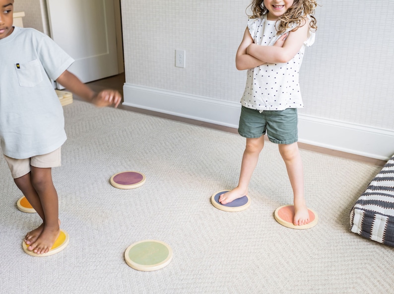A dark-skinned boy with black hair and a light-skinned girl with brown hair playing on the stepping stones in a gray-carpeted living room.