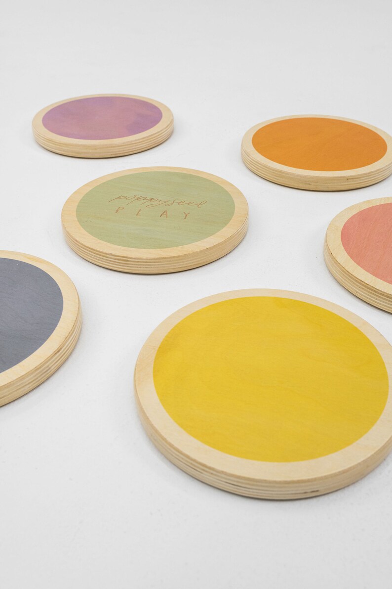 Set of round wooden stepping stones. There are 6 total, one in each of the following colors -- blue, yellow, pink, green, orange, and purple. "Poppyseed PLAY" is engraved on the green stepping stone.