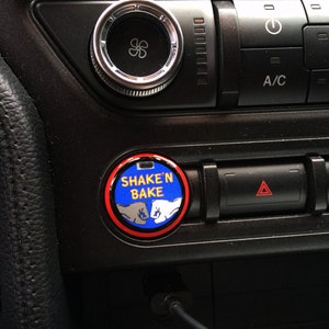 May include: A car's dashboard with a round button that says "SHAKE'N BAKE" in white text on a blue background, with two fist-bumping hands. The button is surrounded by a red ring. Other buttons and a vent control are also visible.