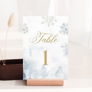 May include: A table number card with a winter forest design. The card features a watercolor illustration of pine trees and snowflakes in shades of blue and gray. The word "Table" and the number "1" are in gold script.