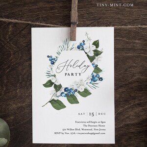 K&ouml;nnte beinhalten: Eine wei&szlig;e Einladungskarte f&uuml;r eine Weihnachtsfeier, gehalten von einer Holzklammer. Die Karte zeigt ein Aquarelldesign eines floralen Kranzes mit blauen Beeren und wei&szlig;en Bl&uuml;ten. Der Text "Holiday PARTY" ist in eleganter Schrift dargestellt.