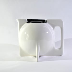 May include: A white ceramic teapot with a square base and a rounded front. The teapot has a black rectangular lid and a handle on the side. The spout is a long, thin, pointed shape.