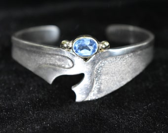 May include: A silver bracelet with a blue gemstone set in a gold setting. The bracelet has a unique, abstract design.