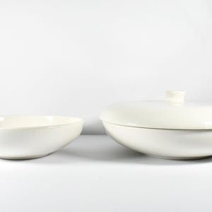 May include: A set of two off-white ceramic serving dishes. One dish is an open, shallow bowl with curved sides. The second dish is a matching lidded casserole with a low-profile, rounded lid featuring a small, centered knob handle on top.