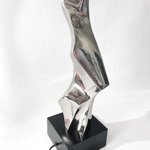 May include: A modern silver-toned table lamp with an abstract, sculptural design. The lamp features a polished metal body with a unique, angular shape, mounted on a black square base. A brown power cord extends from the base.