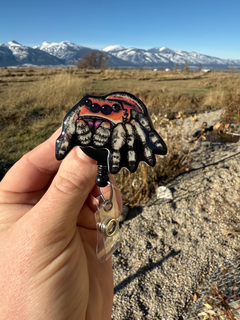 May include: A black and white spider badge reel with a colorful jumping spider design. The badge reel has a retractable cord and a clear plastic ID holder. The spider has orange, red, and pink accents. The background shows a field and mountains.