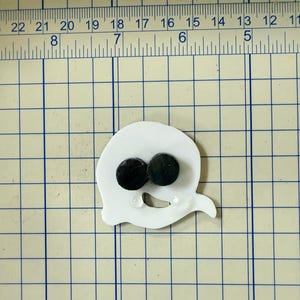 May include: A white ghost-shaped craft element with two black circular eyes. The ghost is positioned on a grid with numbered markings, suggesting a crafting or measuring context. The design is simple and cartoonish.