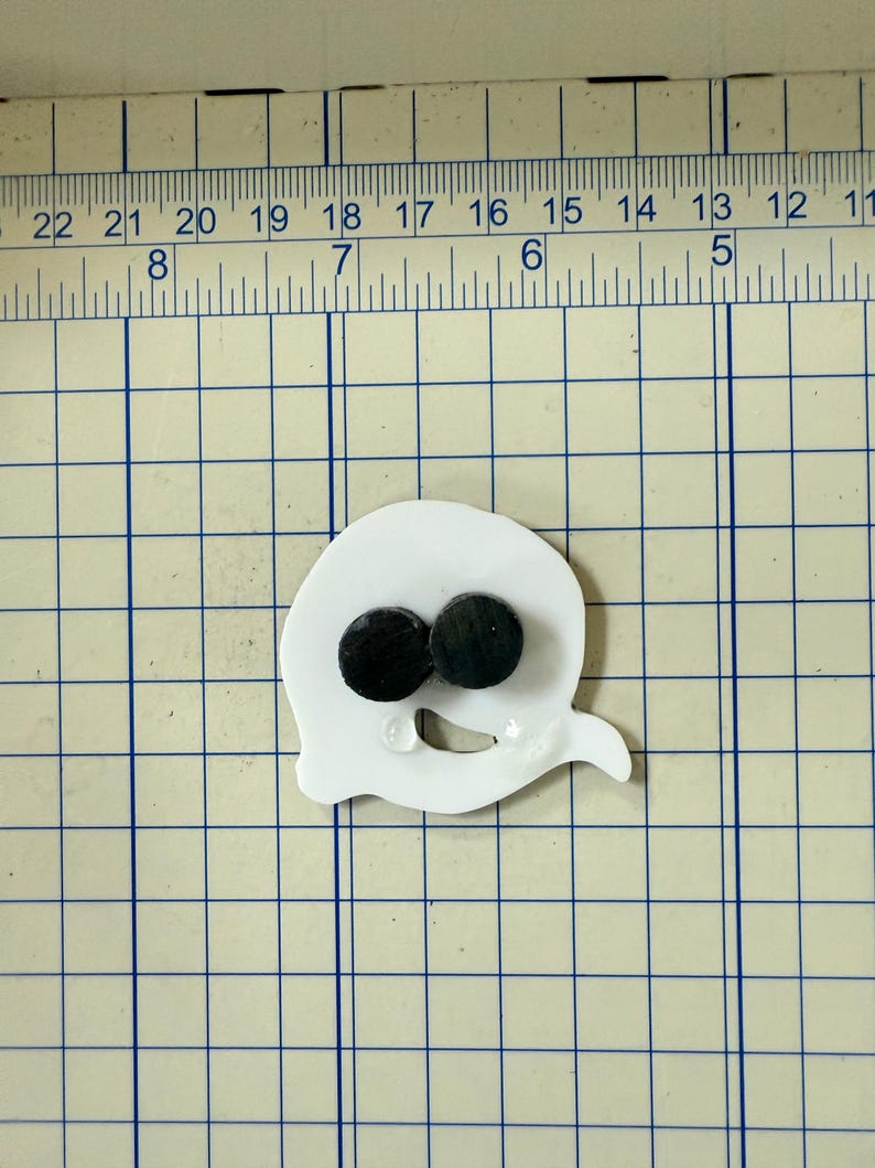May include: A white ghost-shaped craft element with two black circular eyes. The ghost is positioned on a grid with numbered markings, suggesting a crafting or measuring context. The design is simple and cartoonish.