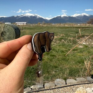 May include: A brown bear badge reel with a black retractable cord. The reel is held in a hand with a mountain range in the background.