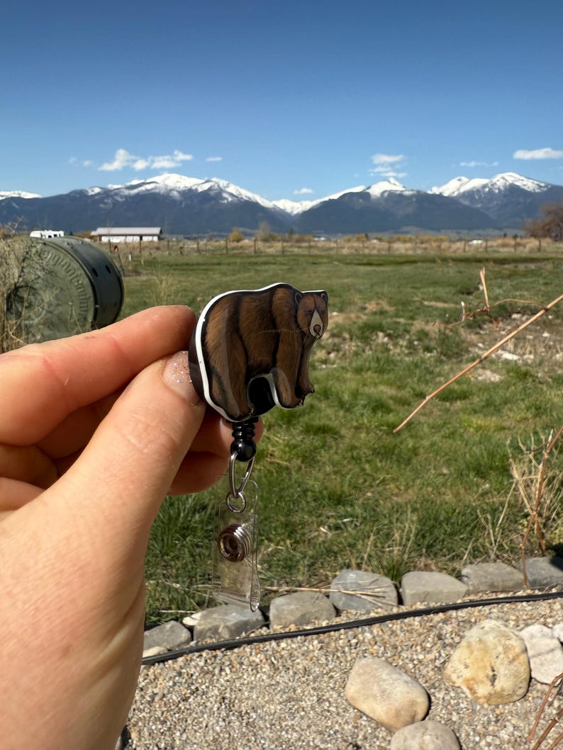 May include: A brown bear badge reel with a black retractable cord. The reel is held in a hand with a mountain range in the background.