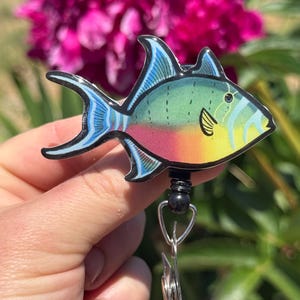 May include: A colorful fish-shaped badge reel with a vibrant design. The fish features shades of green, blue, yellow, and pink. The badge reel has a black bead and a metal clip. The background includes a blurred pink flower.