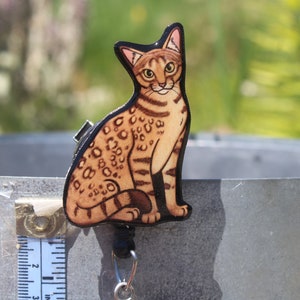 May include: A brown and black wooden cat charm with a black retractable badge reel. The cat is sitting with its tail curled around its legs. The charm is attached to a silver metal chain.