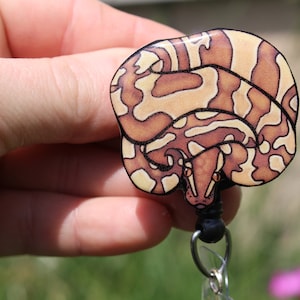 May include: A brown and white patterned snake curled up on a black retractable badge reel. The snake has a black outline and red eyes.