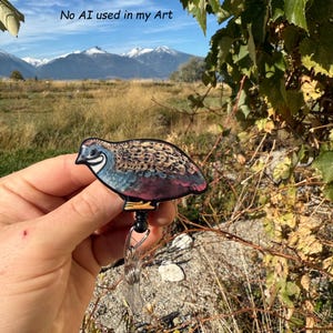 May include: A colorful quail badge reel with a bird design. The badge reel has a black retractable cord and a clear plastic clip. The text "No AI used in my Art" is visible in the background.