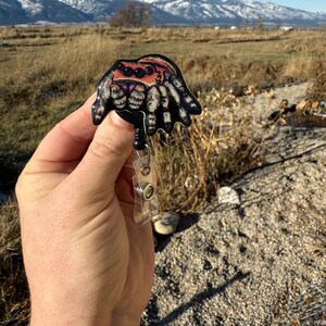 May include: A colorful spider badge reel with a black retractable cord and a clear plastic clip. The spider has a detailed design with orange, pink, and black accents. The background shows a scenic view of mountains and a field.