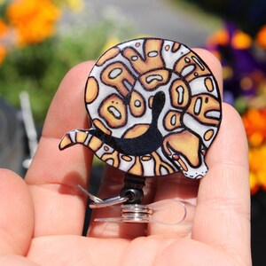 May include: A black, white, and orange patterned reptile-shaped badge reel. The reel is attached to a clear plastic retractable cord.