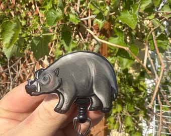 May include: A gray and black badge reel featuring a stylized illustration of a rhino. The rhino has a white snout and is set against a black background. The reel has a retractable cord and a metal clip for attaching to clothing or accessories.