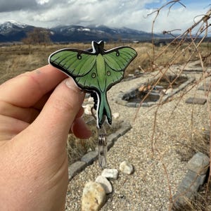 Puede incluir: Un llavero de polilla luna verde y marr&oacute;n con un clip plateado. El llavero se sostiene en una mano contra un fondo de un camino rocoso y monta&ntilde;as en la distancia.
