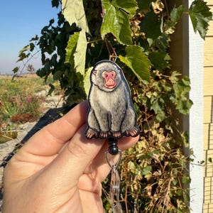 May include: A gray and pink Japanese macaque badge reel. The badge reel features a cartoon illustration of a Japanese macaque with a pink face and gray fur. The reel has a black retractable cord and a clear plastic clip.
