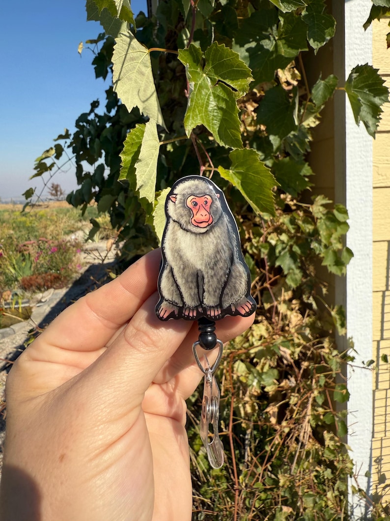 May include: A gray and pink Japanese macaque badge reel. The badge reel features a cartoon illustration of a Japanese macaque with a pink face and gray fur. The reel has a black retractable cord and a clear plastic clip.
