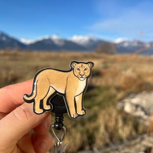 May include: A brown and black cartoon cougar badge reel with a black swivel clip and a clear plastic retractable cord.