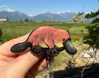 May include: A badge reel featuring a cartoon platypus with orange, yellow, and black coloring. The platypus design is attached to a black retractable reel with a clear plastic clip. The background shows a scenic view of mountains and a blue sky.