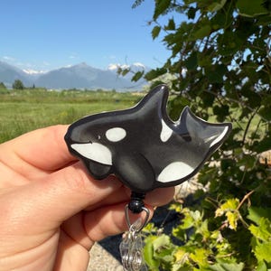 May include: A black and white orca whale retractable badge reel. The orca design has white accents and is attached to a black reel with a clear plastic clip. The background shows a scenic view of mountains and greenery.