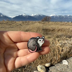 May include: A small, round opossum pin with a black and gray body, pink nose and tail, and white face. The pin is held in a hand against a backdrop of mountains and a cloudy sky. Text in the image reads "No AI used in my Art."