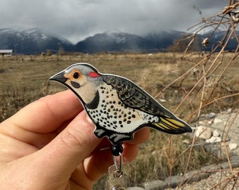 May include: A black and white bird with red accents on its head, printed on a badge reel. The bird is perched on a branch with its wings spread. The badge reel is made of metal and has a clear plastic holder.