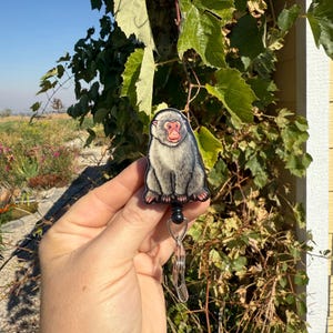 May include: A badge reel featuring a Japanese macaque, also known as a snow monkey. The badge has a gray body, a red face, and is attached to a retractable reel with a metal clip. The background includes green foliage and a blue sky.