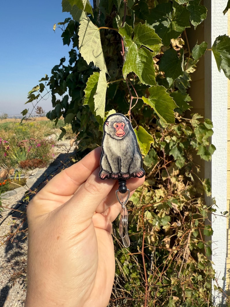 May include: A badge reel featuring a Japanese macaque, also known as a snow monkey. The badge has a gray body, a red face, and is attached to a retractable reel with a metal clip. The background includes green foliage and a blue sky.