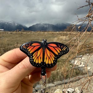 May include: A black and orange butterfly-shaped badge reel with a clear plastic retractable cord. The butterfly has black wings with orange and yellow markings.