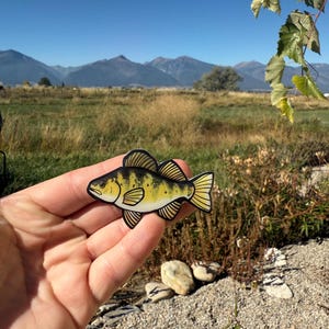 May include: A yellow perch fish-shaped sticker with black stripes and accents. The sticker is held in front of a scenic background of mountains, fields, and a clear blue sky. The fish sticker is a detailed illustration.