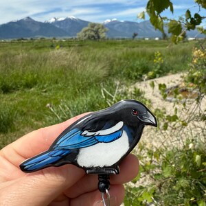 May include: A badge reel featuring a glossy, illustrated magpie bird with black, white, and vibrant blue plumage. The bird has a small red eye and a black beak. The badge reel mechanism is attached to the underside of the bird figure.