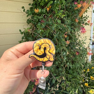 May include: A circular badge reel featuring a yellow and black patterned snake illustration. The snake is coiled in a circular shape with a black stripe running along its body. The badge reel has a clear plastic strap and a metal clip mechanism.